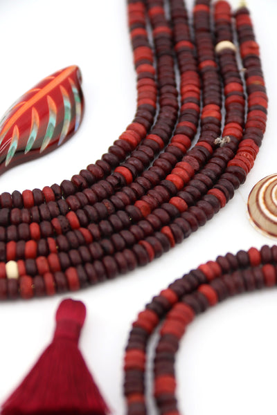 Flatlay of burgundy and rustic red bone heishi beads in strands, handmade and ethically sourced, styled with tassel and decorative accents.