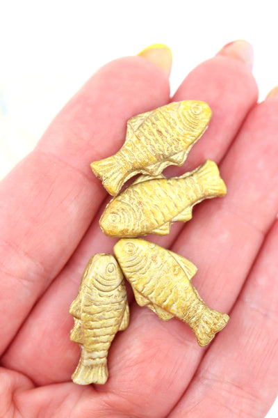 Hand holding four matte gold Czech glass fish beads with textured scale detail and a rustic, aged finish.

