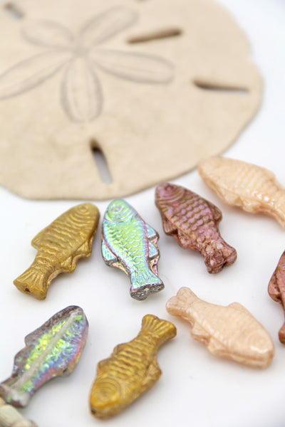 Assorted Czech glass pressed fish beads in gold, cream, mauve, and vitrail finishes arranged around a sand dollar on a white background.

