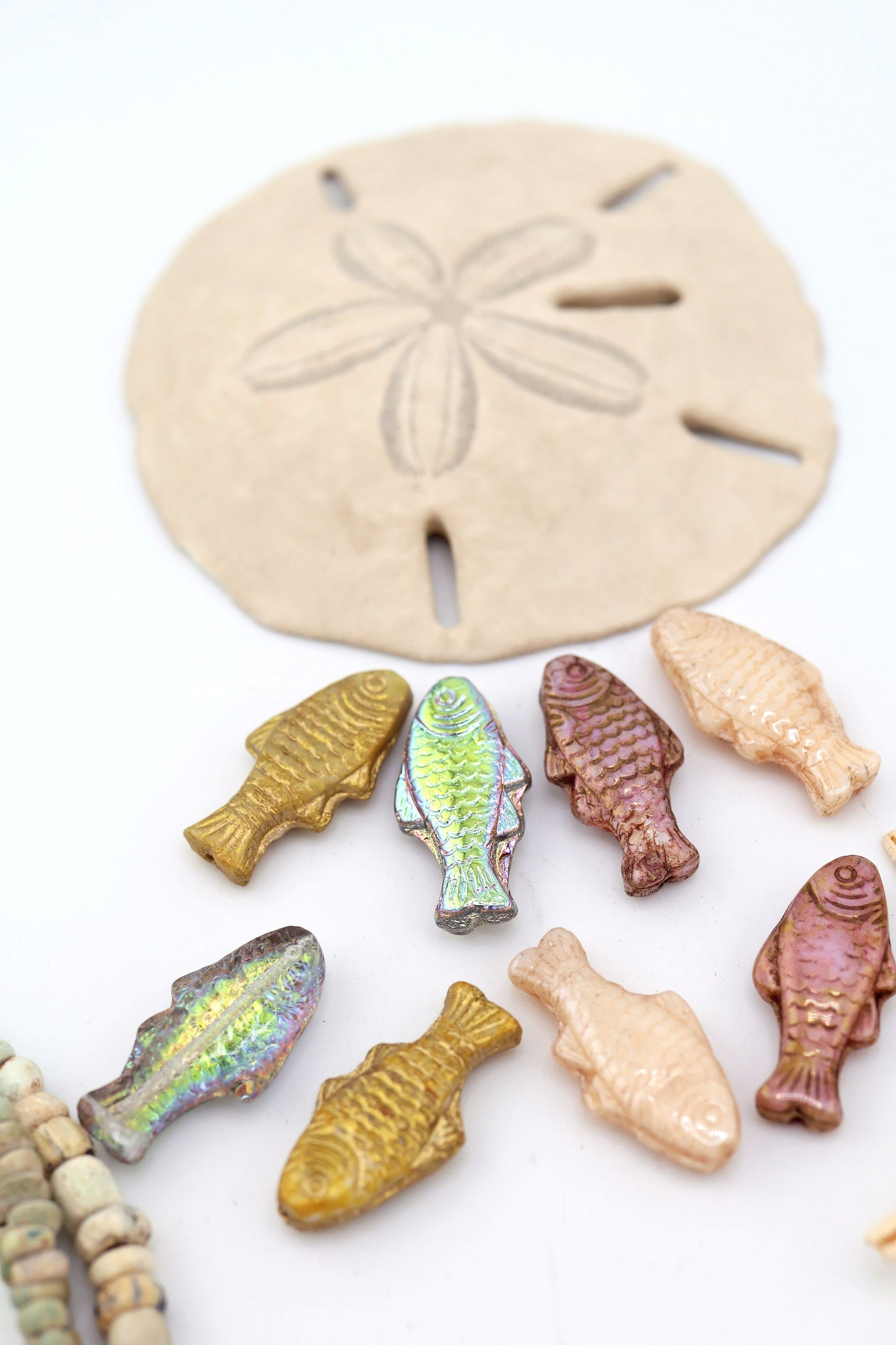 Assorted Czech glass pressed fish beads in gold, cream, mauve, and vitrail finishes arranged around a sand dollar on a white background.

