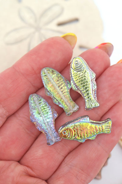 Hand holding four iridescent vitrail Czech glass fish beads in shimmering hues of lime, aqua, and lavender.

