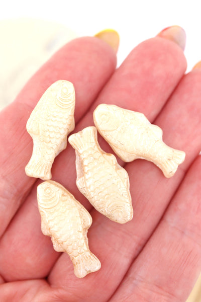 Hand holding four creamy ivory Czech glass fish beads with subtle gold wash and engraved scale detailing.

