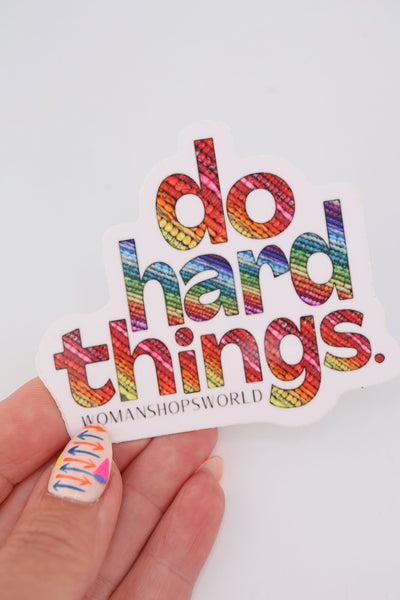 Close-up of a rainbow vinyl sticker that reads “do hard things.” Each bold letter is filled with colorful bead textures. The sticker has a white border and the WomanShopsWorld logo underneath.