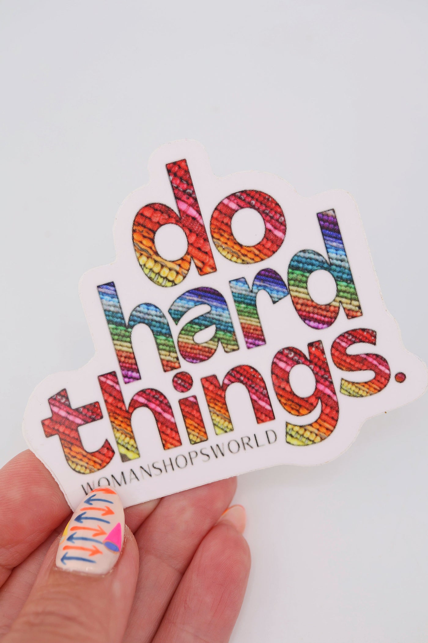 Close-up of a rainbow vinyl sticker that reads “do hard things.” Each bold letter is filled with colorful bead textures. The sticker has a white border and the WomanShopsWorld logo underneath.