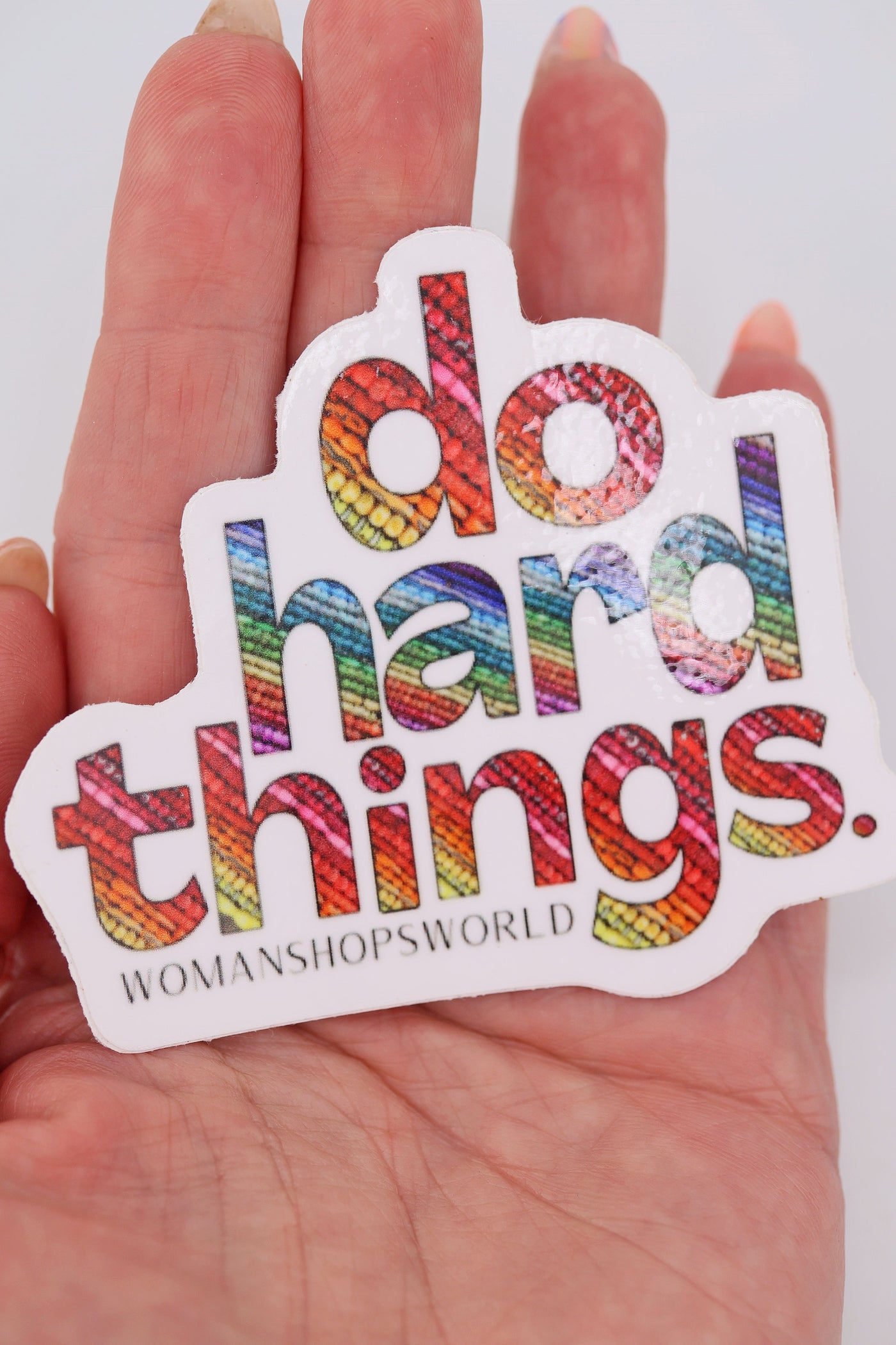 Close-up of a rainbow vinyl sticker that reads “do hard things.” Each bold letter is filled with colorful bead textures. The sticker has a white border and the WomanShopsWorld logo underneath.