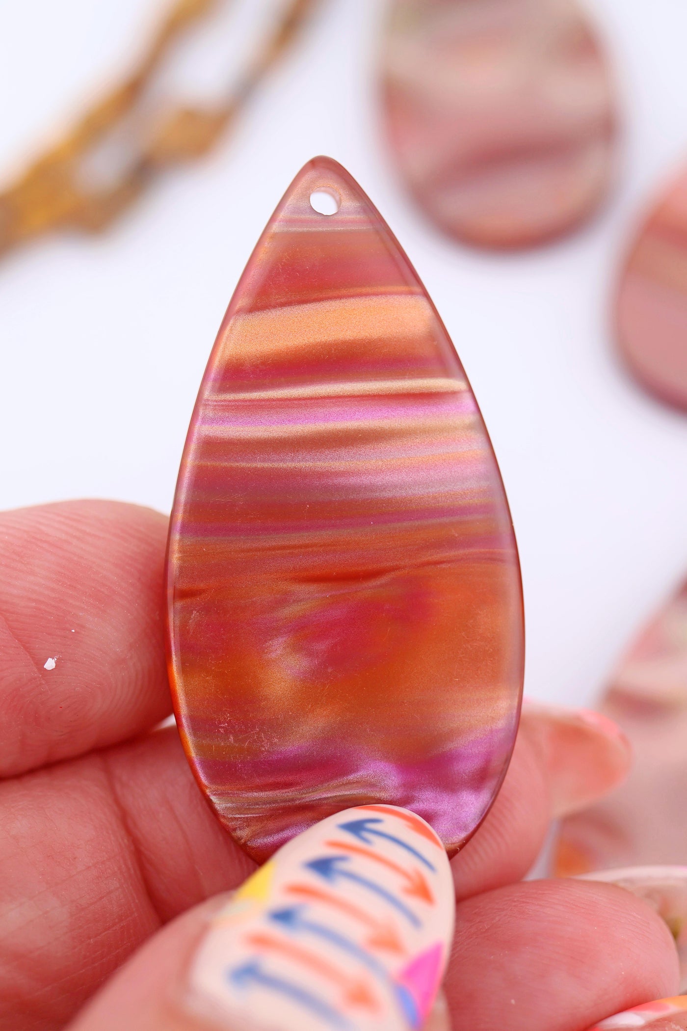 Hand holding a single Italian resin teardrop pendant showing glowing stripes of peach, pink, and copper with a pearlescent finish.