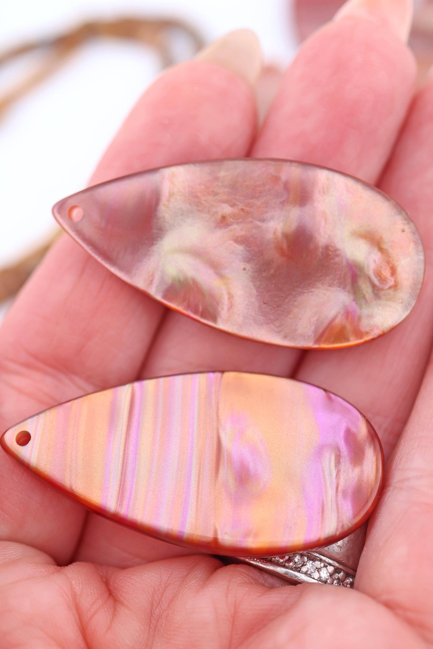 Hand holding two Italian resin teardrop pendant showing glowing stripes of peach, pink, and copper with a pearlescent finish.