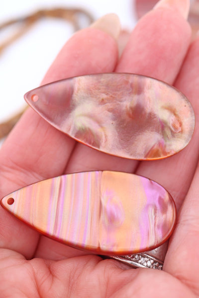 Hand holding two Italian resin teardrop pendant showing glowing stripes of peach, pink, and copper with a pearlescent finish.