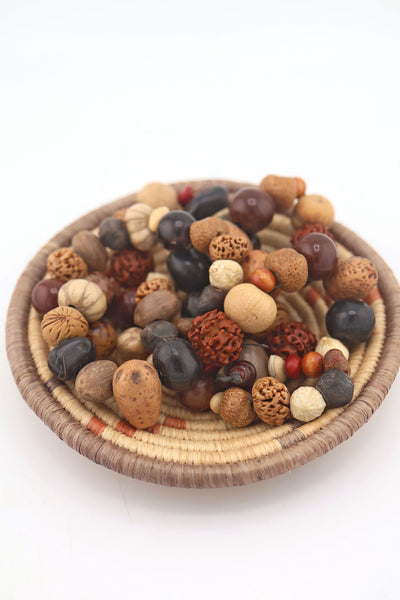Strand of assorted natural nut and seed beads in earthy brown, tan, and black tones, featuring rudraksha and palm nuts, in a woven basket.