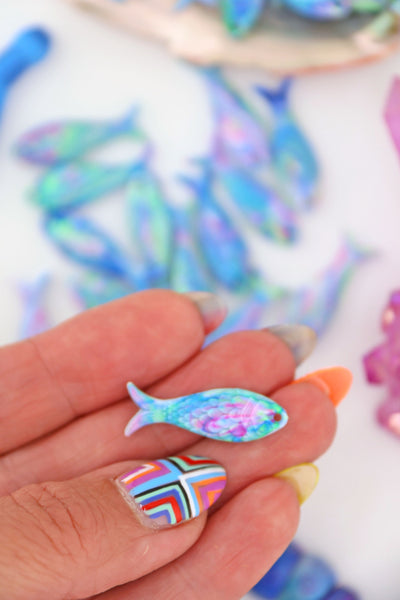 Hand holding a Pastel Aurora resin fish charm with iridescent blue, pink, and green swirls, scale texture visible.