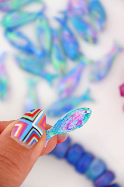 Hand holding a Pastel Aurora resin fish charm with iridescent blue, pink, and green swirls, scale texture visible.