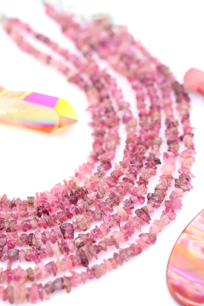 Close-up of natural pink tourmaline chip beads in shades of blush and raspberry, arranged in soft strands beside an iridescent crystal point.