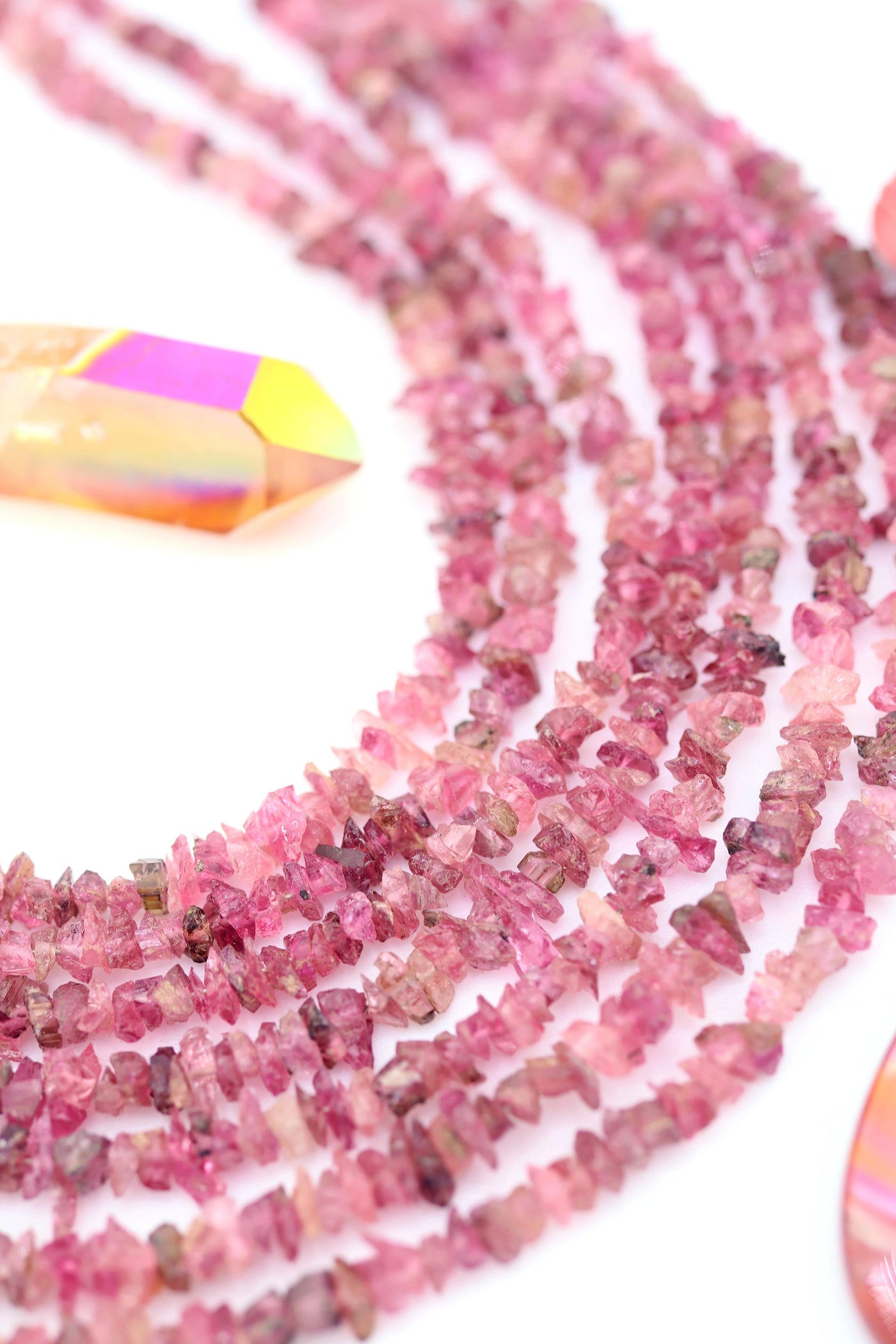 Close-up of natural pink tourmaline chip beads in shades of blush and raspberry, arranged in soft strands beside an iridescent crystal point.