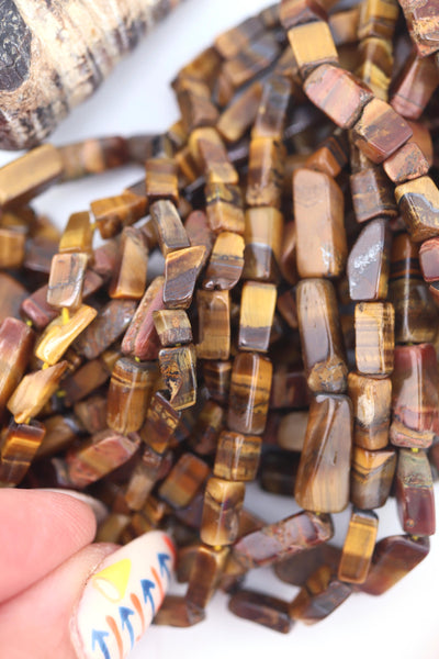 Close-up of natural Tiger’s Eye gemstone beads in rectangular box shape, showing golden-brown shimmer and striped detail, styled with a rust tassel.